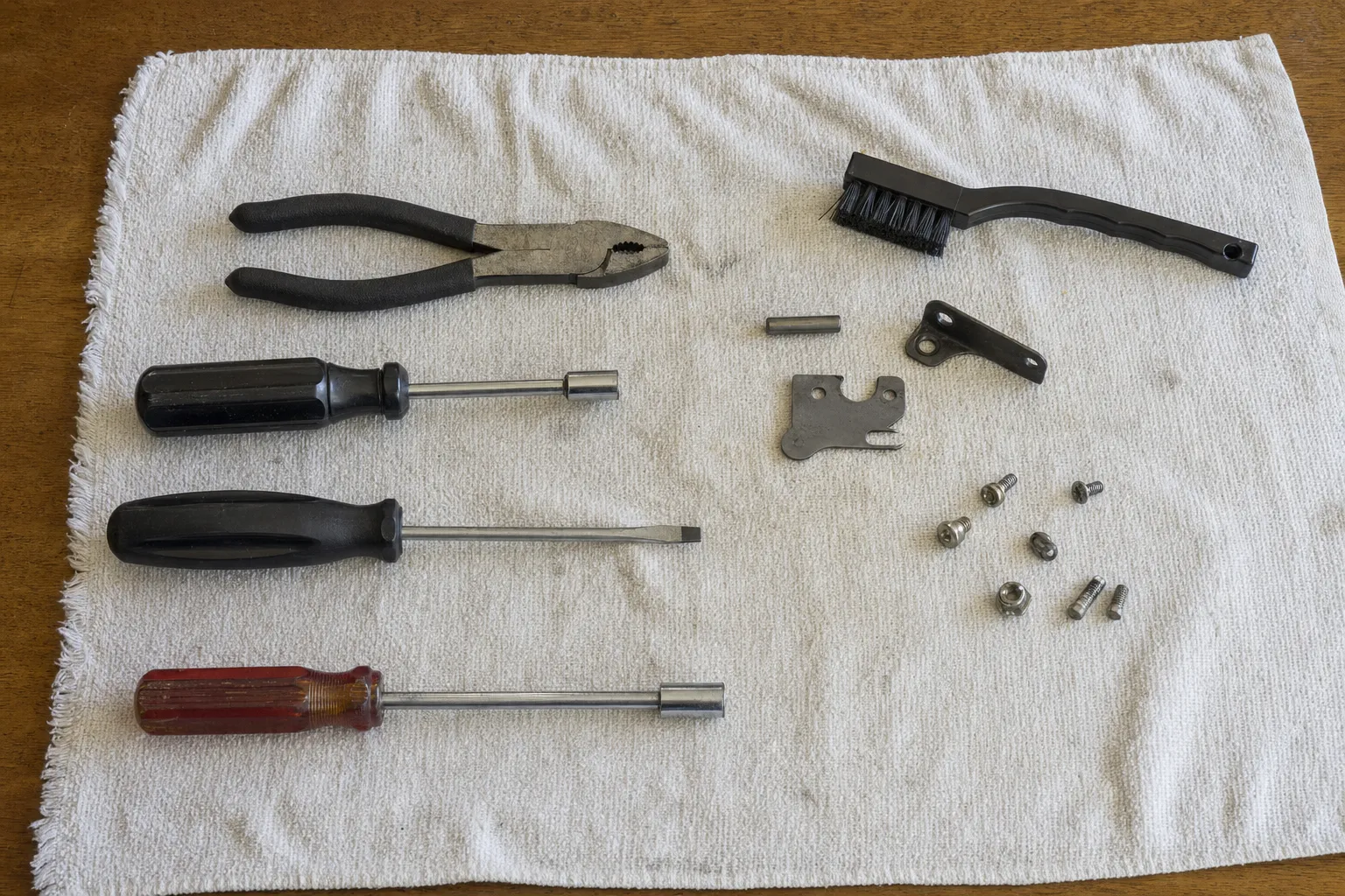 Pliers, two nut drivers and a flat screwdriver on a white towel beside a nylon brush, a notched steel plate, a pin and a few screws